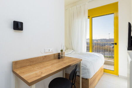 halu! Modern minimalist bedroom featuring a cozy bed and wooden desk adjacent to a bright window with yellow frame allowing natural sunlight to illuminate the space
