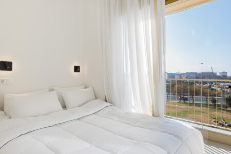 Halu! Bright and airy bedroom with modern decor featuring a cozy white bed and large window offering a scenic view perfect for relaxation and morning sunlight.