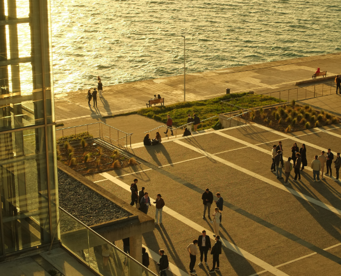 People walking and relaxing on the waterfront promenade near Megaro Mousikis Thessalonikis, with golden sunlight reflecting off the sea.