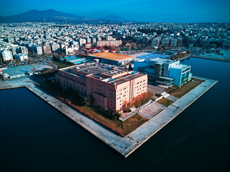 Aerial view of the two contrasting buildings of Megaro Mousikis Thessalonikis—one brown-brick and the other modern white and glass—situated on the waterfront with the city in the background.