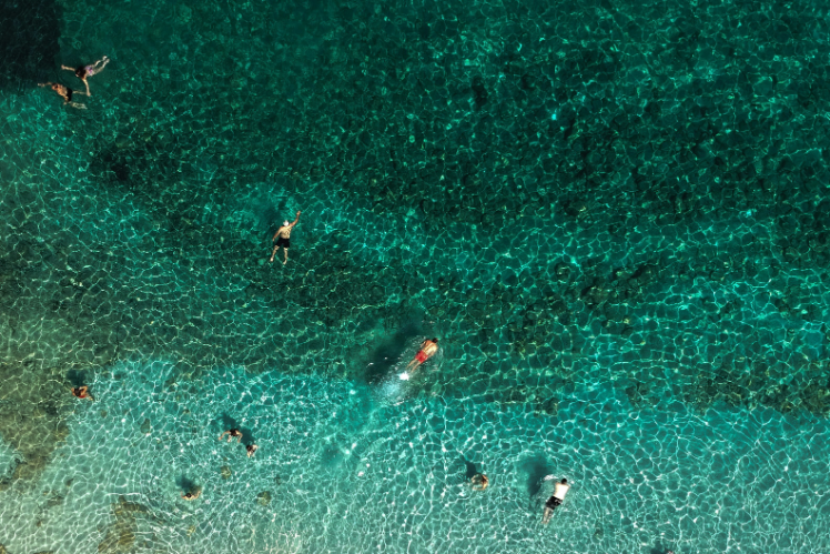 Overhead view of people swimming in the crystal-clear, turquoise waters at a beach in Sivota, highlighting the vibrant shades of blue and green in the water.