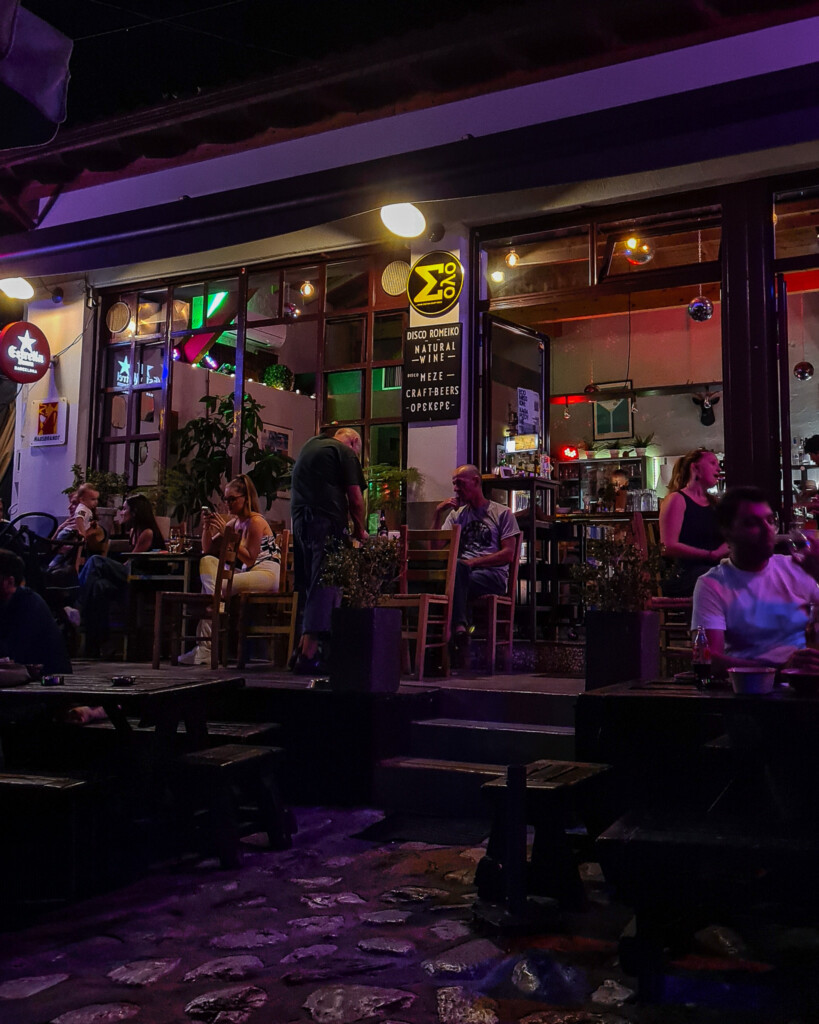 People enjoying drinks at Disco Romeiko bar in Litochoro at night with colorful lights and cozy outdoor seating.