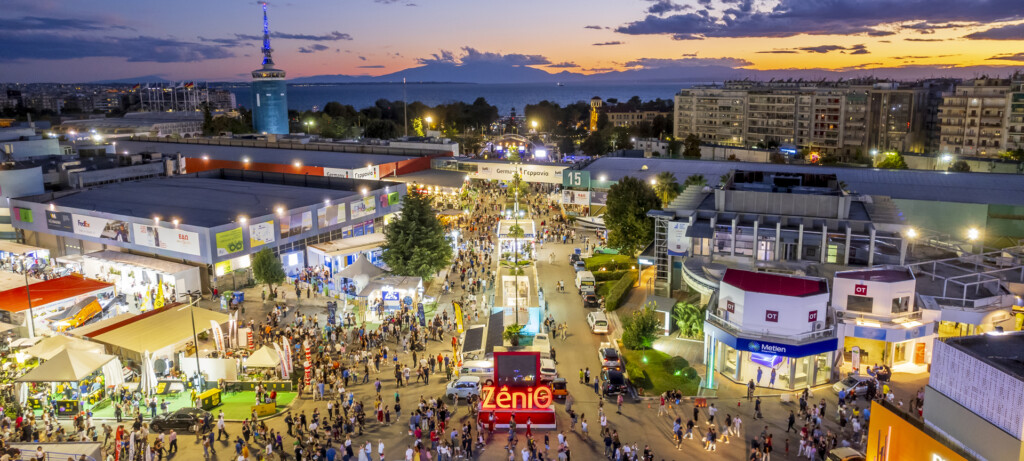Aerial view of Thessaloniki International Fair with exhibitions, crowds, and cityscape at sunset.