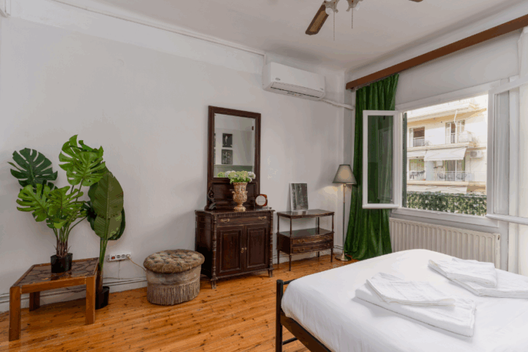 halu! Cozy bedroom with wooden floor features a neatly made bed with white linens a window with natural light a mirror and indoor plants for a refreshing ambiance