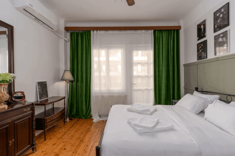 halu! Cozy bedroom with a double bed adorned with white linens and pillows. Room features wooden flooring, green curtains, wall art, and a small desk with a lamp.