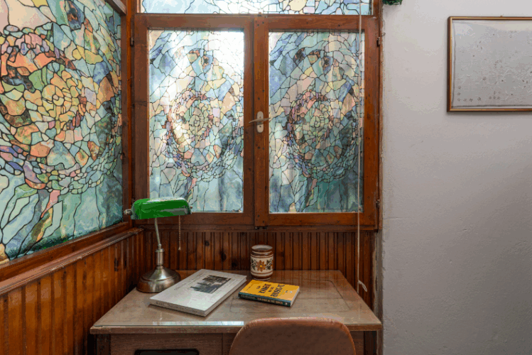 halu! Cozy study nook featuring a wooden desk with books and a green lamp by a window with colorful stained glass creating an inspiring and artistic atmosphere