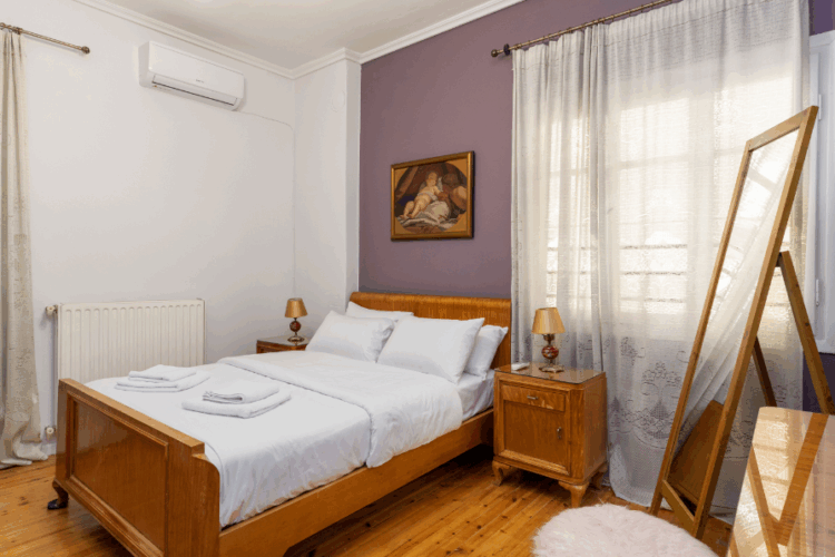 halu! Cozy bedroom featuring a wooden bed with white linens next to a window with curtains a wall painting and a standing mirror on a polished wooden floor.