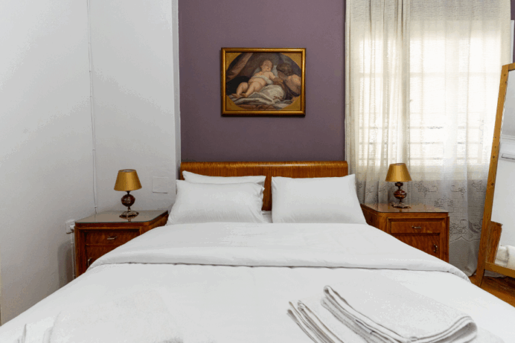 halu! Cozy bedroom with a luxurious white bedding set and elegant framed artwork above the bed. Soft natural light filters through sheer curtains creating a peaceful ambiance