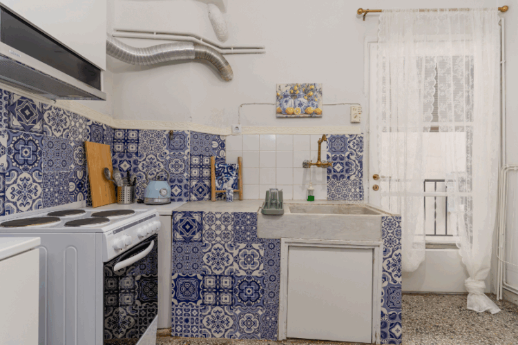 Halu! A cozy kitchen with vintage blue patterned tiles and modern appliances, featuring a stove, sink, and window with sheer curtains letting in natural light.