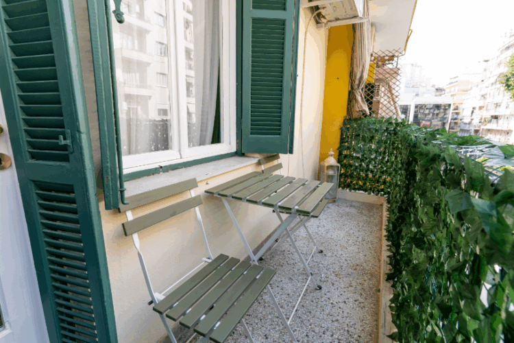 Halu! Cozy balcony with green shuttered windows and a small table set ideal for relaxation. The railing is adorned with lush greenery adding a touch of nature.