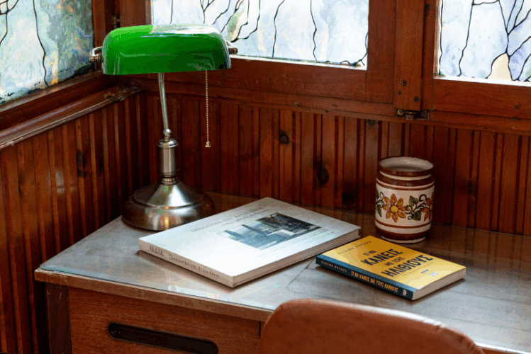 halu! Cozy reading nook with a classic green desk lamp a hardcover book a notebook with a bright yellow cover and a floral ceramic mug on a wooden table near stained glass windows