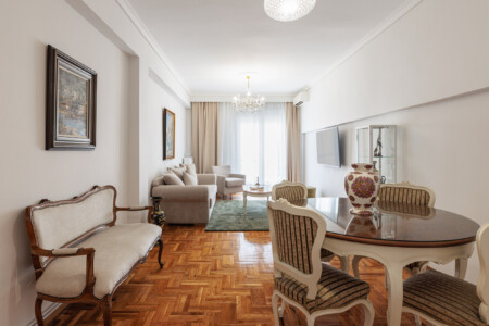 Elegant living room with classical décor, cozy sofa and armchairs, flat-screen TV, and a dining area. Large windows bring natural light, creating a bright and welcoming atmosphere.