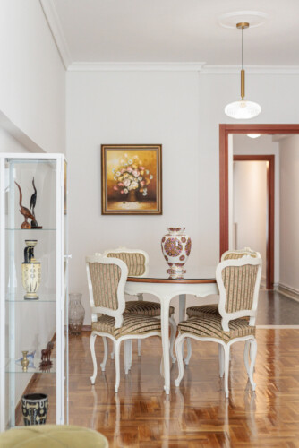 Elegant living room with classical décor, cozy sofa and armchairs, flat-screen TV, and a dining area. Large windows bring natural light, creating a bright and welcoming atmosphere.