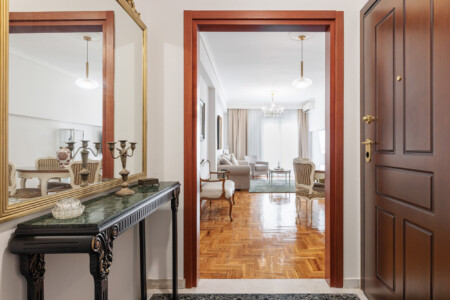 Elegant living room with classical décor, cozy sofa and armchairs, flat-screen TV, and a dining area. Large windows bring natural light, creating a bright and welcoming atmosphere.