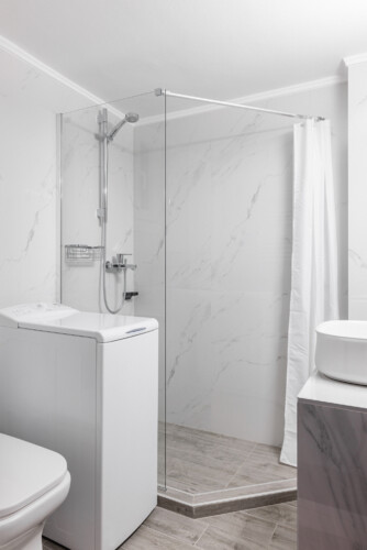 Modern bathroom with elegant marble-style tiles, a glass shower, fresh towels, and a washing machine. Bright and stylish, designed to offer both comfort and practicality during your stay.