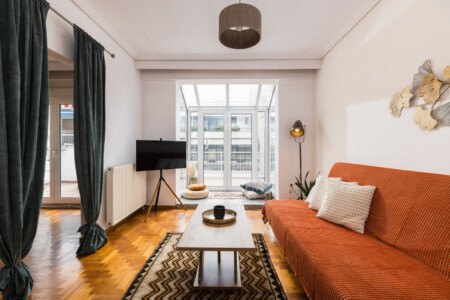 halu! Bright and cozy living room with a modern design featuring an orange sofa a wooden coffee table and a large window providing natural light and a city view