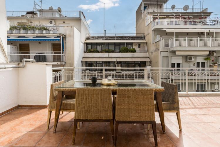 Halu! Cozy urban balcony view with wicker dining set under clear sky. Surrounded by buildings the peaceful setting is perfect for relaxation or outdoor meals.
