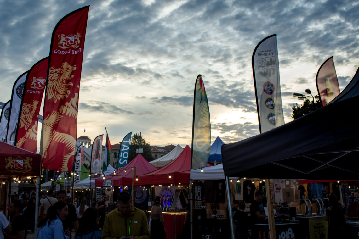 Halu! A bustling outdoor market with colorful tents and flags under a cloudy sky. People enjoy the vibrant atmosphere and diverse stalls at this lively event.