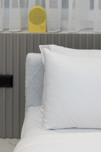 halu! Cozy bedroom with a neatly made bed featuring white pillows and a soft duvet against a modern headboard. A yellow speaker adds a pop of color on the windowsill.