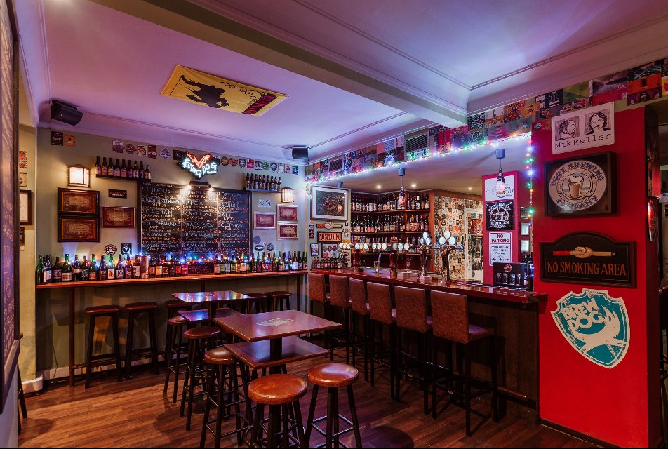 Halu! Cozy bar interior featuring wooden stools and tables, a well-stocked bar, ambient lighting, and vibrant decor creating a warm and inviting atmosphere.