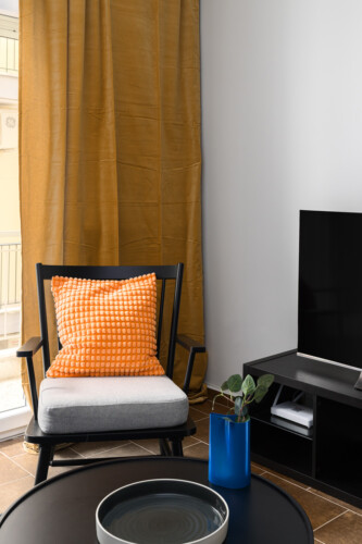 Halu! Cozy living room corner with a black chair featuring a bright orange cushion a flat-screen TV and a sleek gray table with a blue vase and green plant nearby.