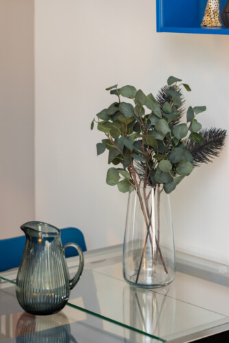 Halu! A stylish glass vase with eucalyptus branches sits on a modern glass table beside a ribbed glass jug creating a minimalist and elegant home decor scene.