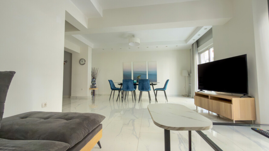 Halu! Modern living room with sleek furniture including a large TV and dining area with blue chairs. Bright natural lighting enhances the spacious and stylish decor.