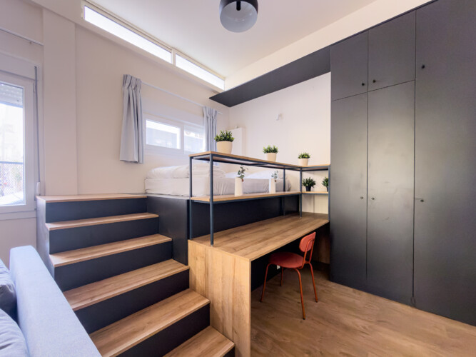 halu! Modern studio apartment with a stylish loft design featuring a wooden staircase leading to a raised sleeping area with sleek black storage units and a workspace.