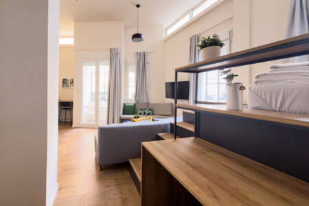 halu! Bright and modern living room with wooden shelves and cozy gray sofa. Large windows allow natural light. Stylish decor with plants adds a fresh ambiance.