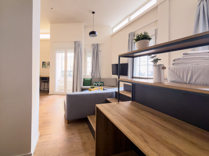 halu! Bright and modern living room with wooden shelves and cozy gray sofa. Large windows allow natural light. Stylish decor with plants adds a fresh ambiance.