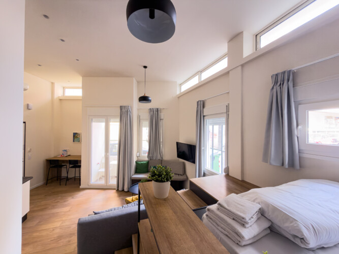 Halu! Bright modern living space with large windows natural light and cozy furnishings featuring a bed sofa small dining area and a stack of fresh towels on the table.