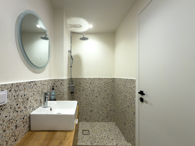 halu! Modern minimalist bathroom featuring a round mirror above a wooden countertop with a white sink and terrazzo tiles providing a stylish and functional design.