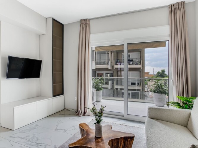 halu! Bright and modern living room with a wall-mounted TV and stylish furniture. Large windows open to a balcony view enhancing natural light and ambiance.