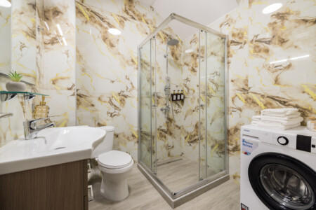 Halu! Experience modern luxury at Ourania Lofts and Suites in Thessaloniki. Elegant bathroom with a glass shower, white sink, washing machine, and marble-style walls.